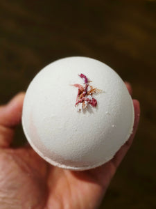 White and pink bath bomb with dried flowers held in a hand against a blurred background.