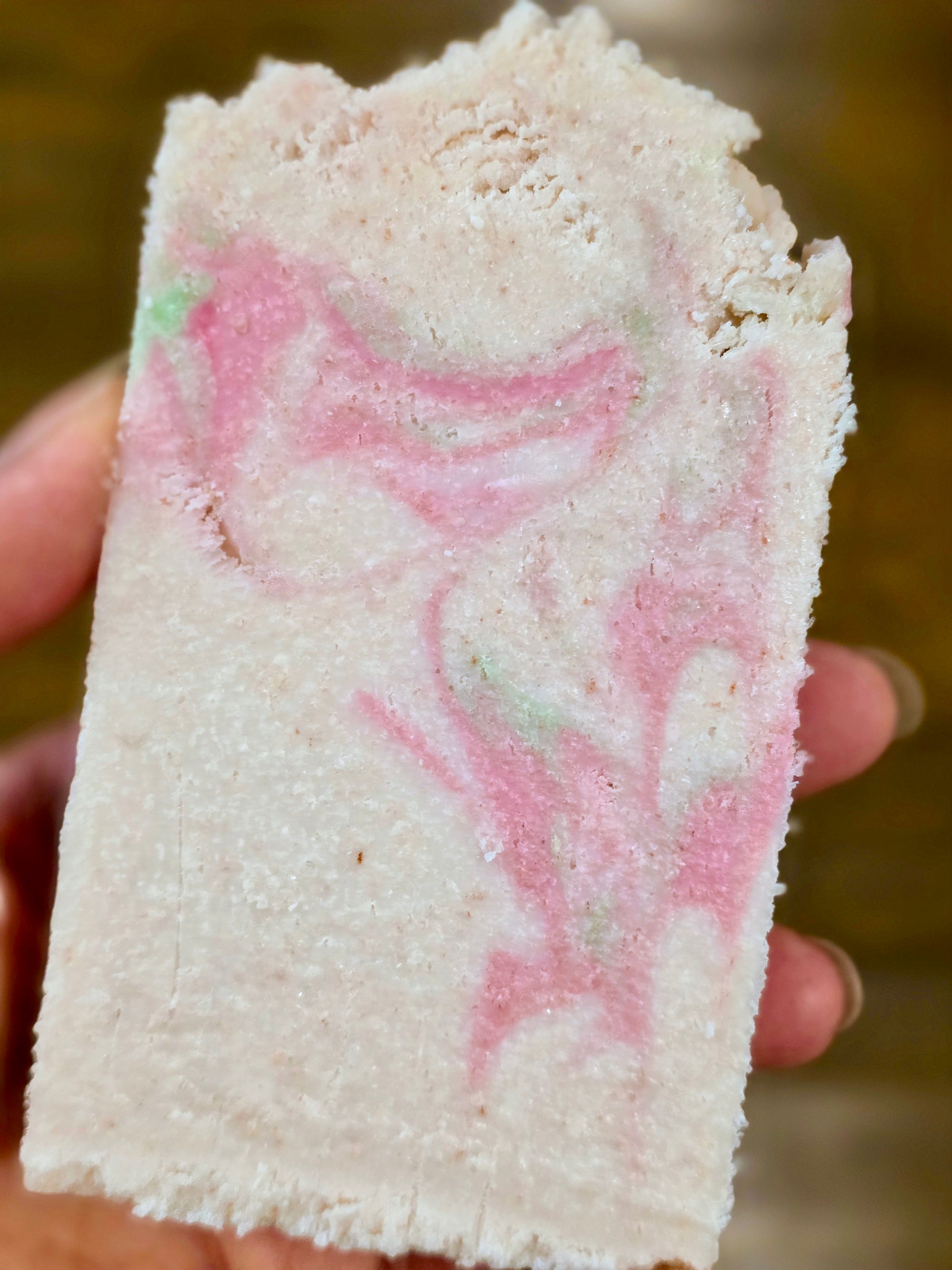 A bar of soap with pink and green swirls held by a hand against a blurred natural background.